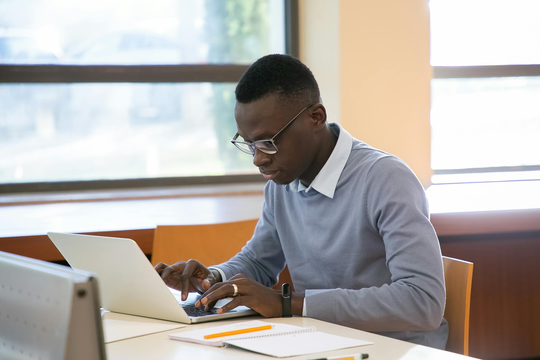 Concentrated young African American male student in formal clothes and eyeglasses reading notes in planner and typing on laptop while doing assignment in modern workspace