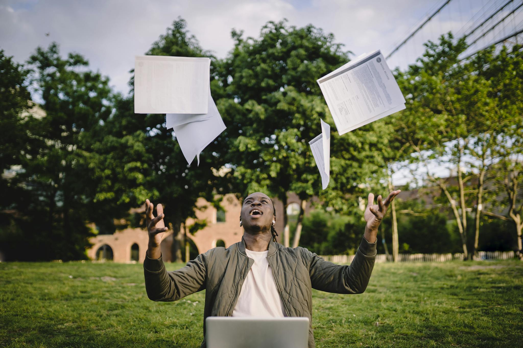 Happy young black male freelancer throwing papers while celebrating successful project during remote work in green park